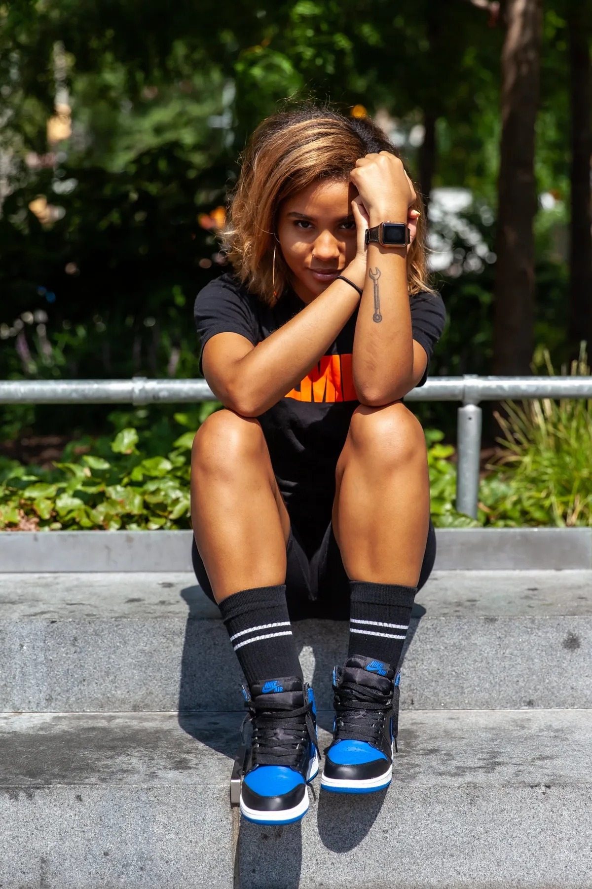 A woman sits on a concrete ledge with her hands in her hair, wearing a black tee and blue-and-black high-top sneakers