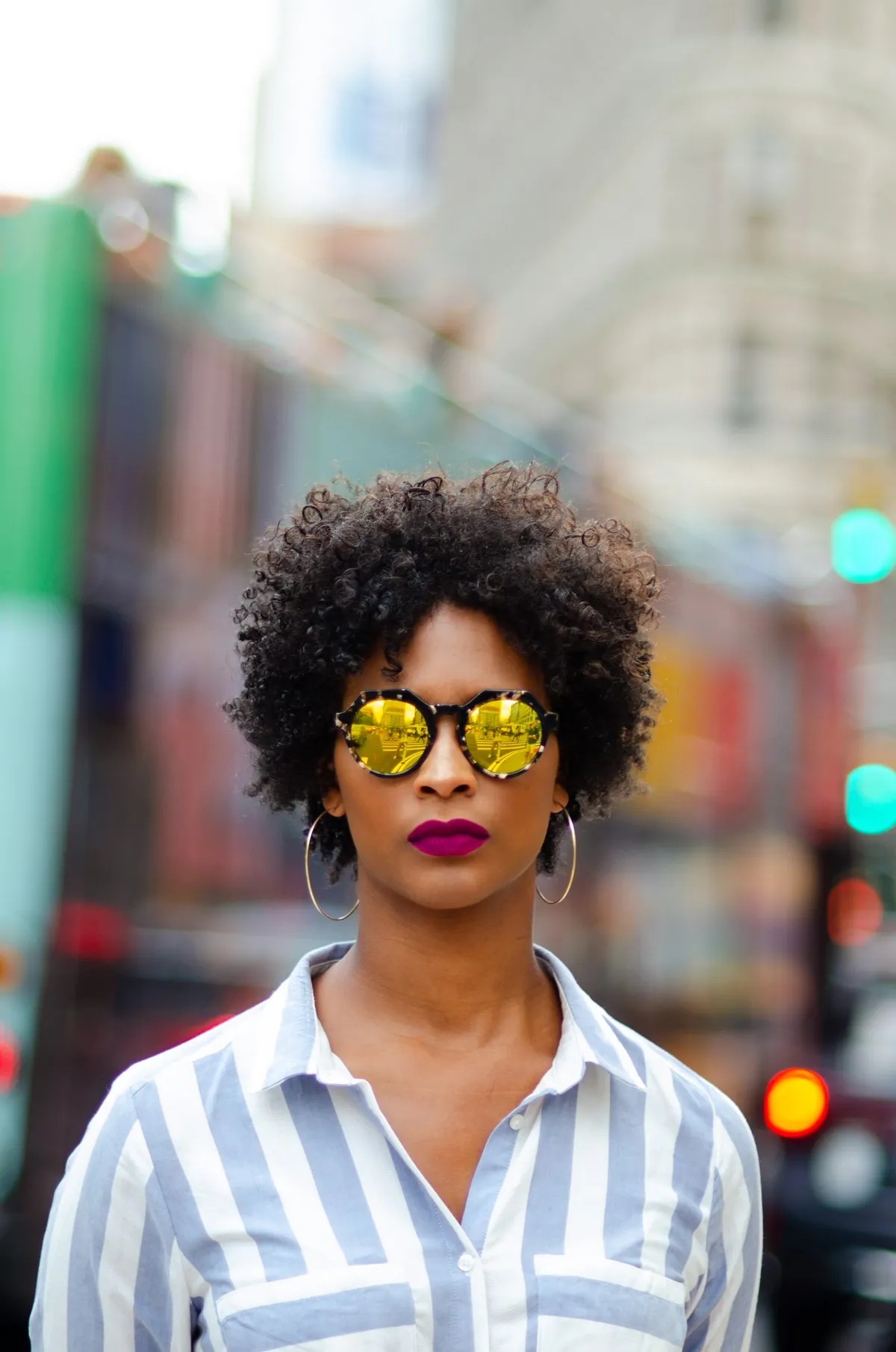A woman with a curly natural afro in mirrored round sunglasses and a striped shirt stands on a busy city street
