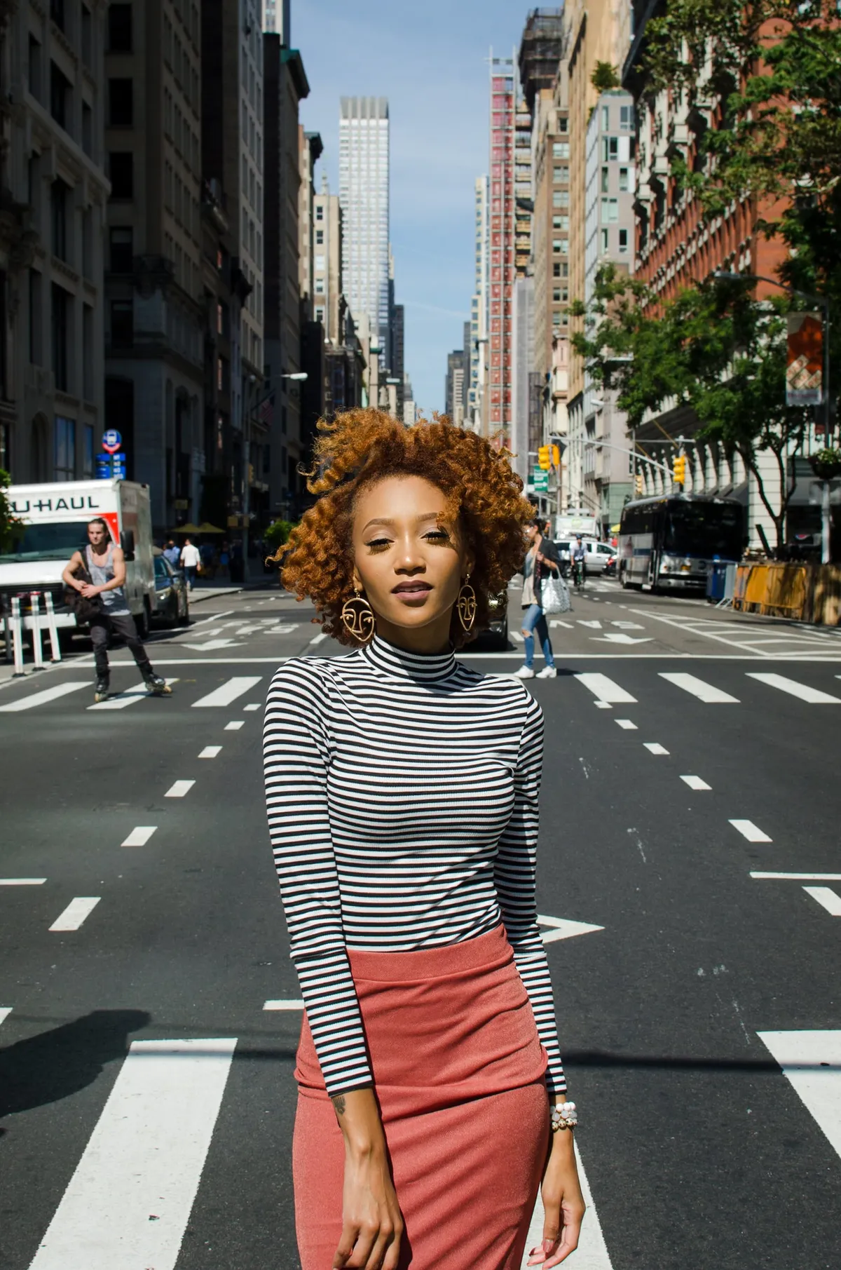 A woman with curly auburn hair in a striped turtleneck and coral skirt stands in the middle of a Manhattan avenue