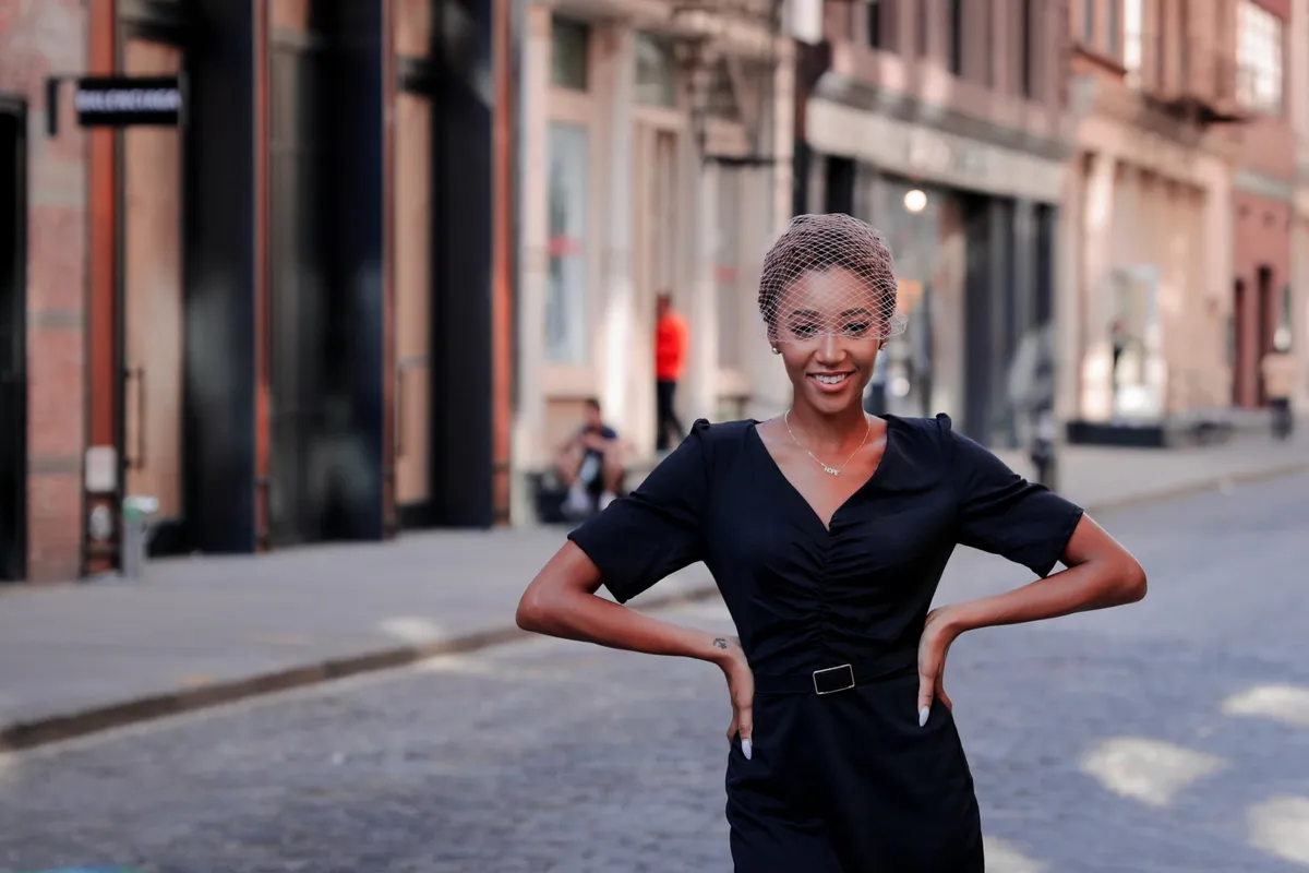 A woman in a black wrap dress poses with her hands on her hips on a SoHo cobblestone street