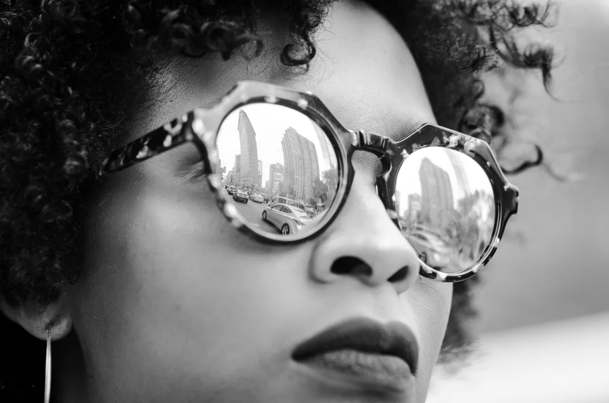 Black-and-white close-up of a woman in round sunglasses reflecting the city skyline