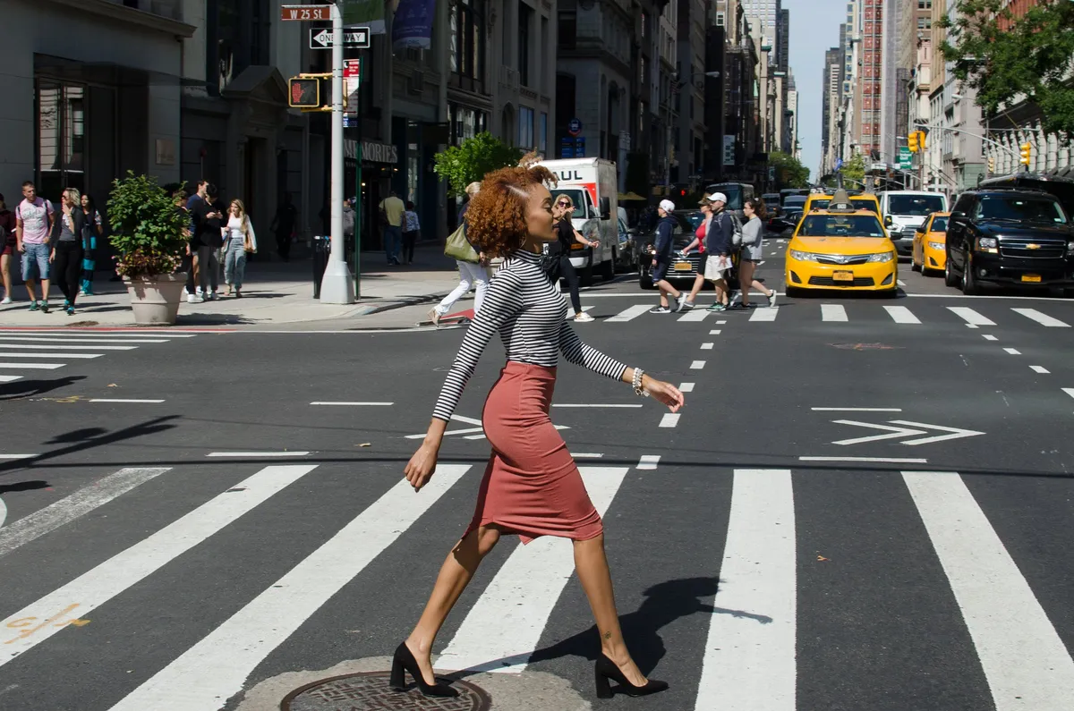 A woman in heels crosses a city crosswalk, yellow taxis lined up behind her