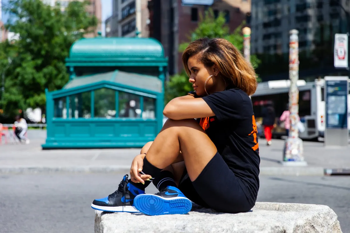 A woman sits on a granite block beside a green subway kiosk, knees drawn up in blue sneakers