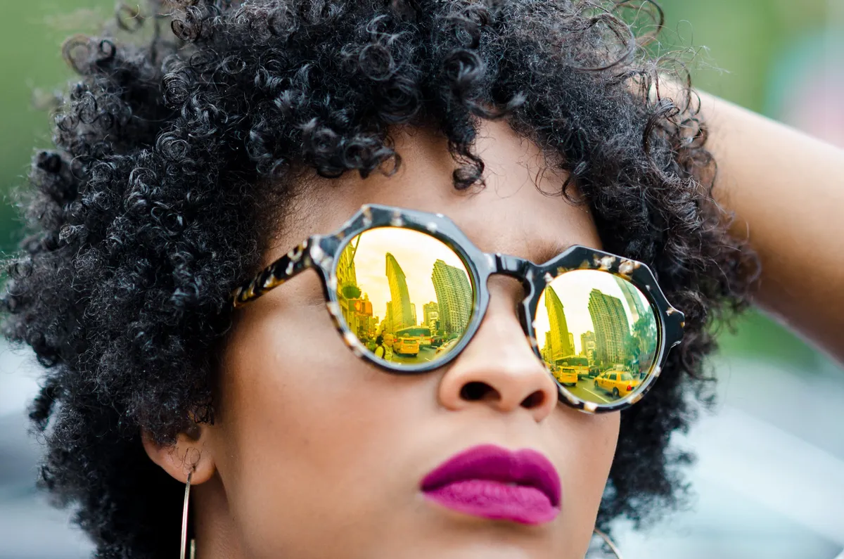 Close-up of a woman with magenta lipstick in gold mirrored sunglasses, hand in her curly hair