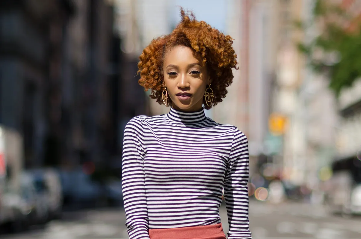 A woman with voluminous auburn curls in a striped turtleneck looks into the camera on a sunlit street