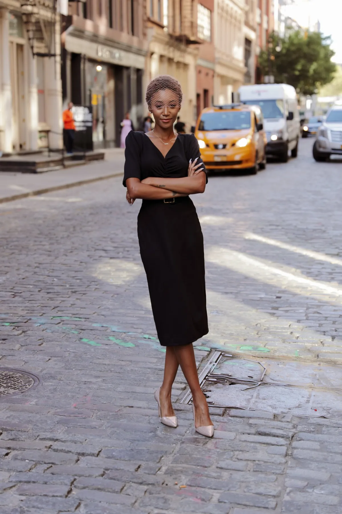 A woman with crossed arms stands on a cobblestone street with yellow taxis behind her