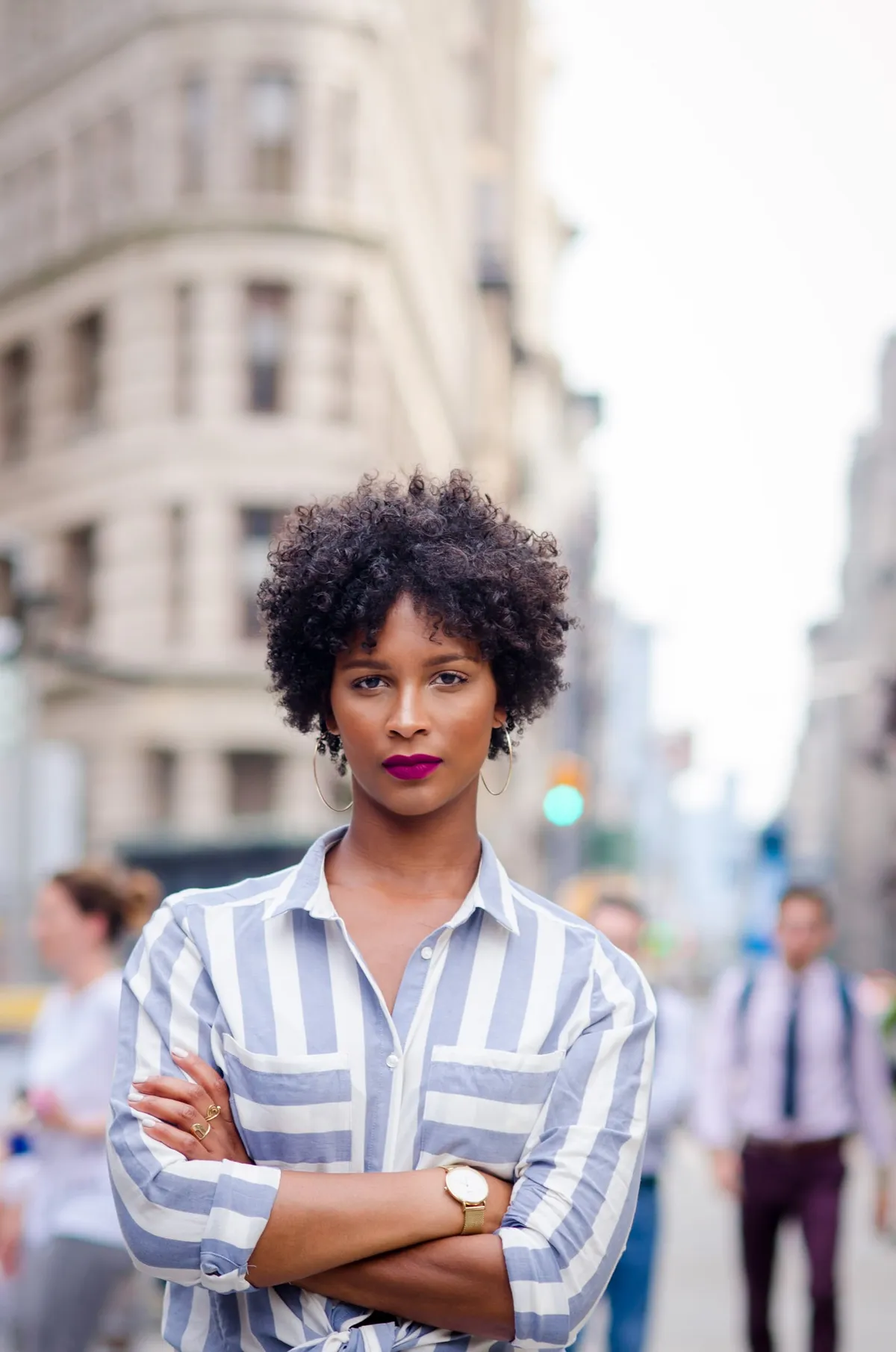 A woman with arms crossed stands on a Manhattan sidewalk in a blue-and-white striped shirt, pedestrians blurred behind