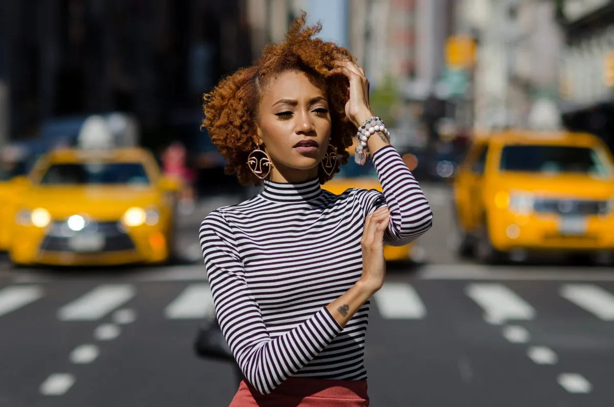 A woman touches her hair while yellow taxis wait at the crosswalk behind her
