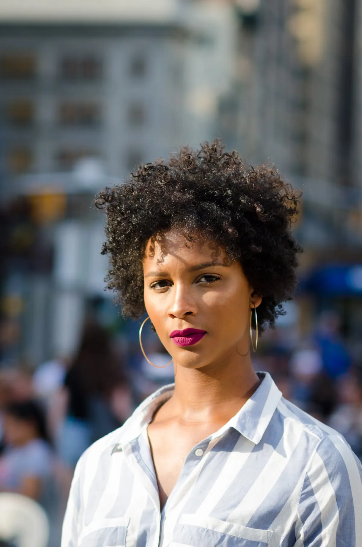 A woman in a striped shirt and hoop earrings looks into the camera with a crowded city street behind her