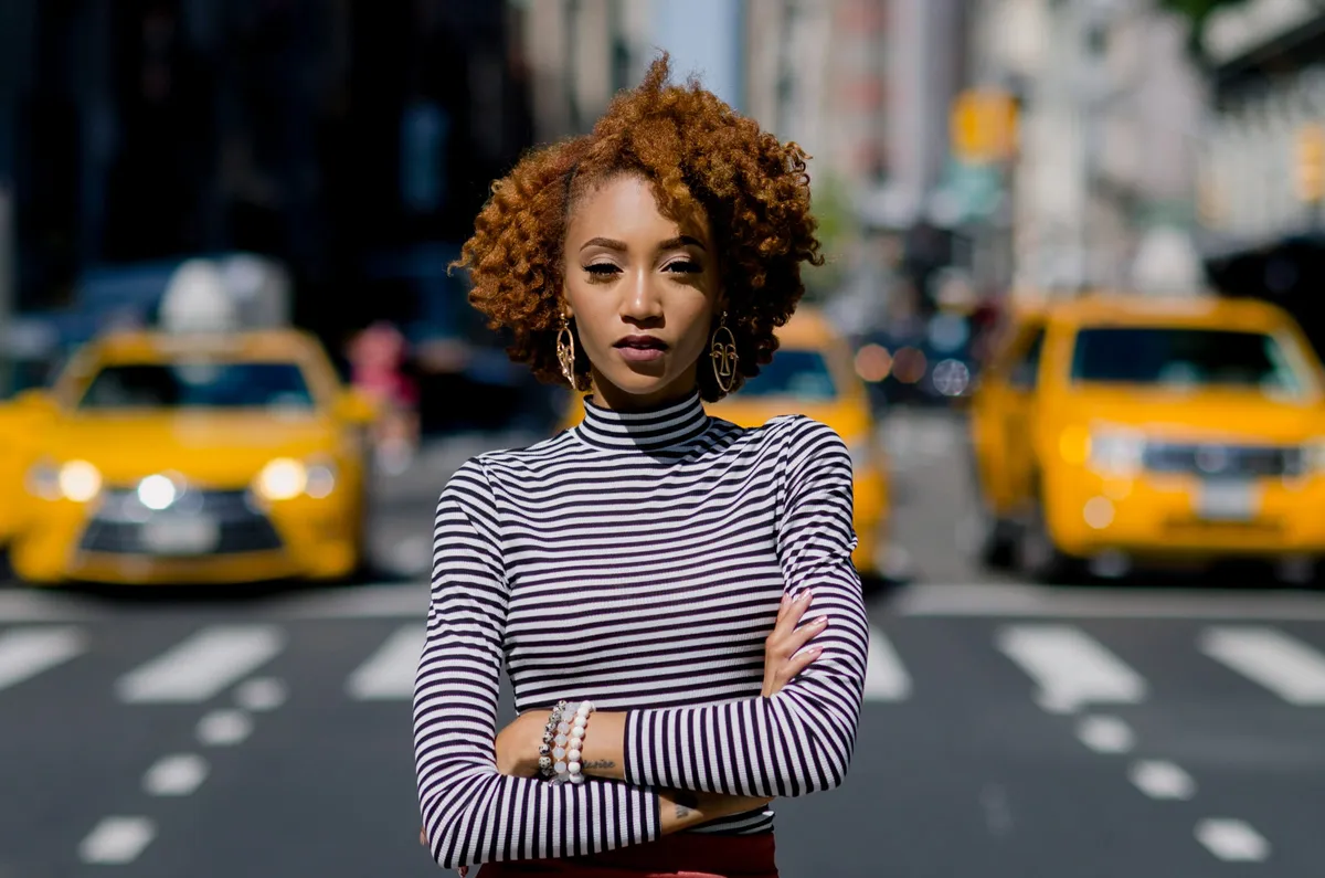 A woman with crossed arms stands in front of a row of blurred yellow taxis
