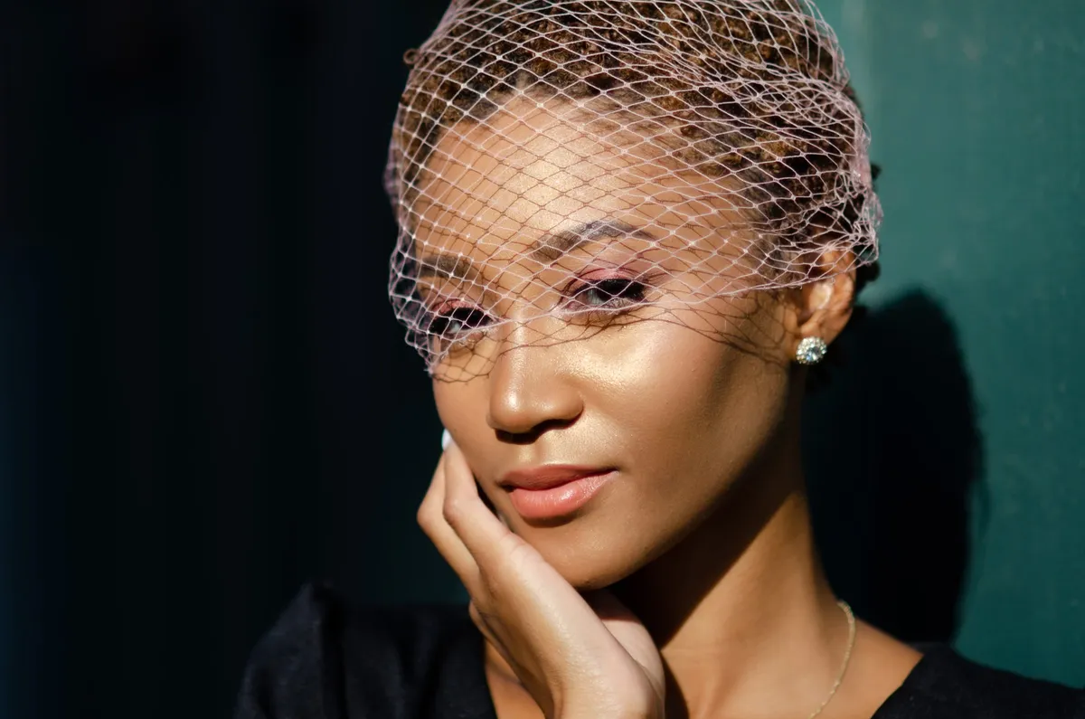 Close-up of a woman in a birdcage veil with her hand at her chin against a teal wall
