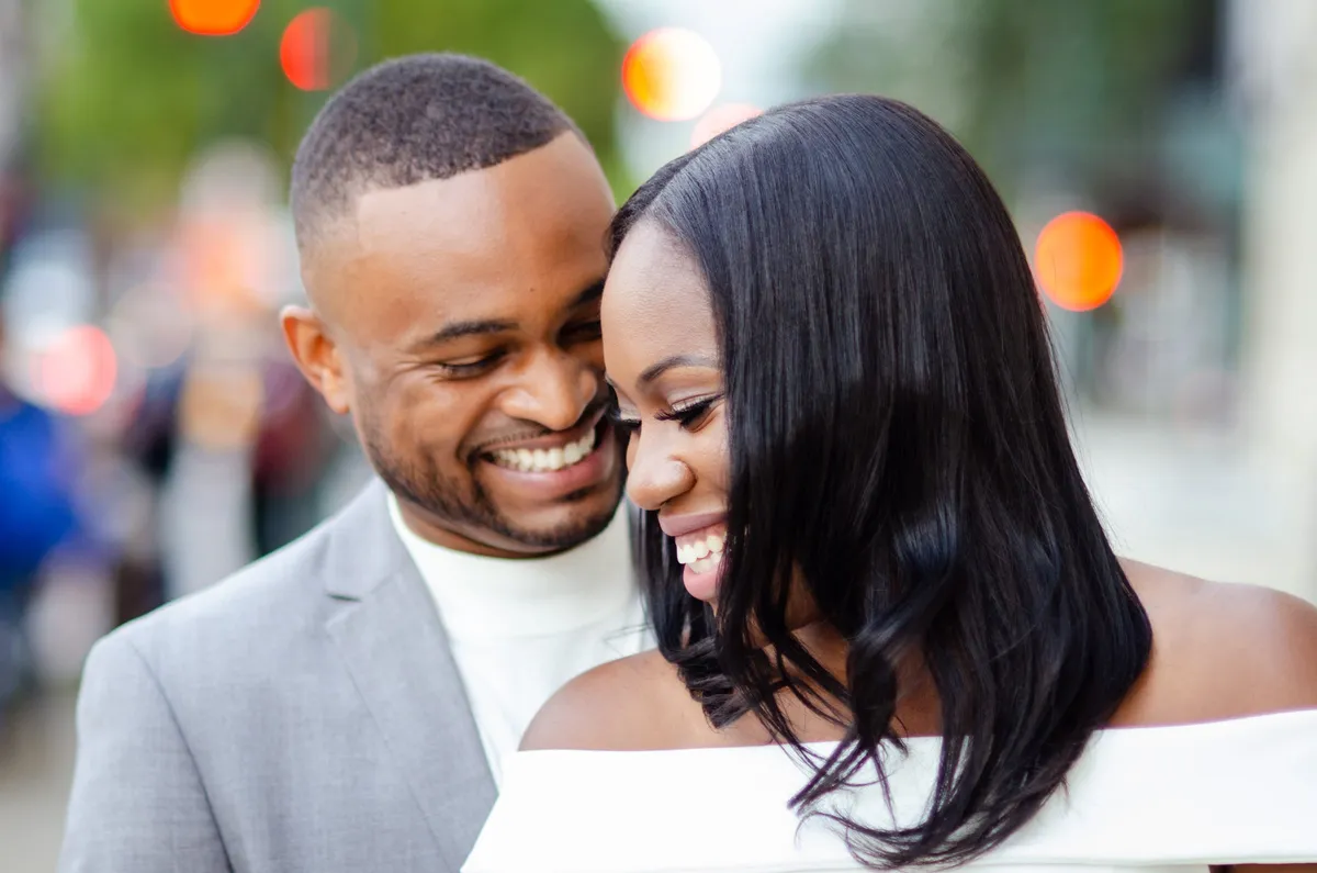 An engaged couple laugh together, cheek to cheek, on a city street at golden hour