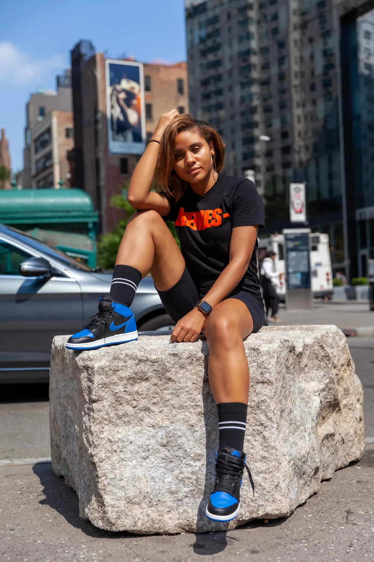 A woman leans back on a granite block with high-rise buildings and a blue sky behind her
