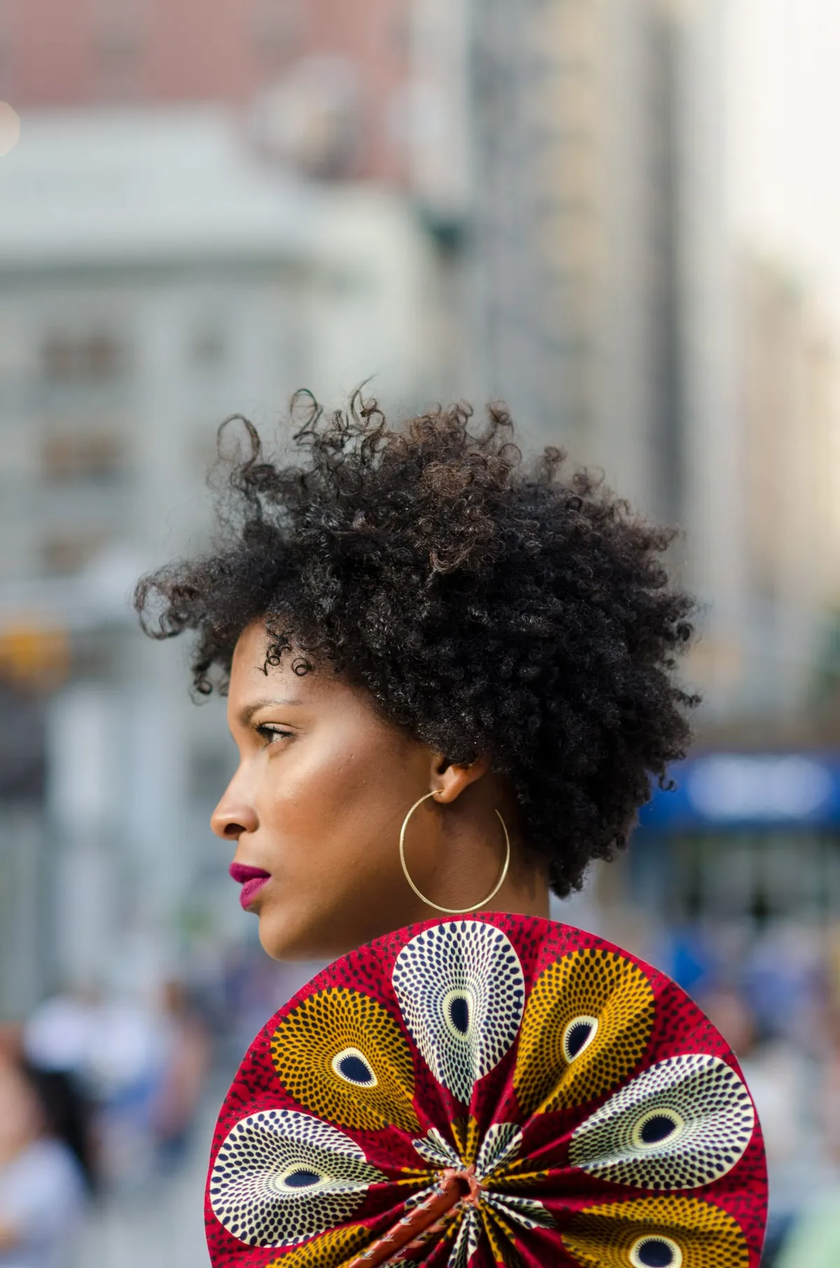 Profile of a woman with a curly afro holding a red and gold patterned fan on a city street