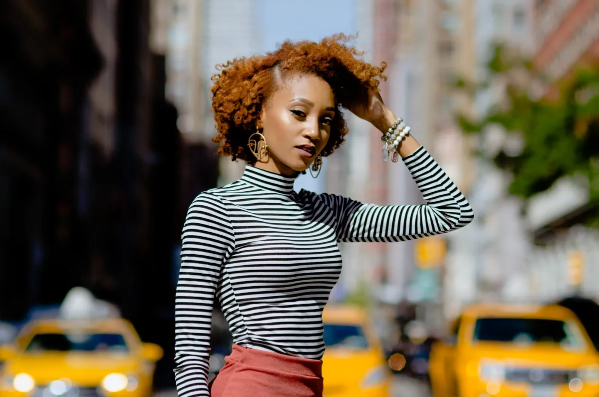 A woman leans into the frame with a hand in her curls, taxis and city buildings behind her