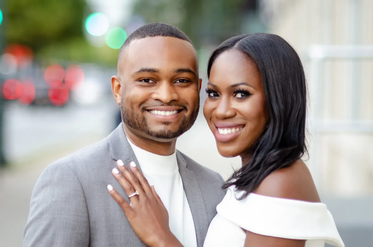 An engaged couple smile at the camera, her hand with an engagement ring resting on his chest