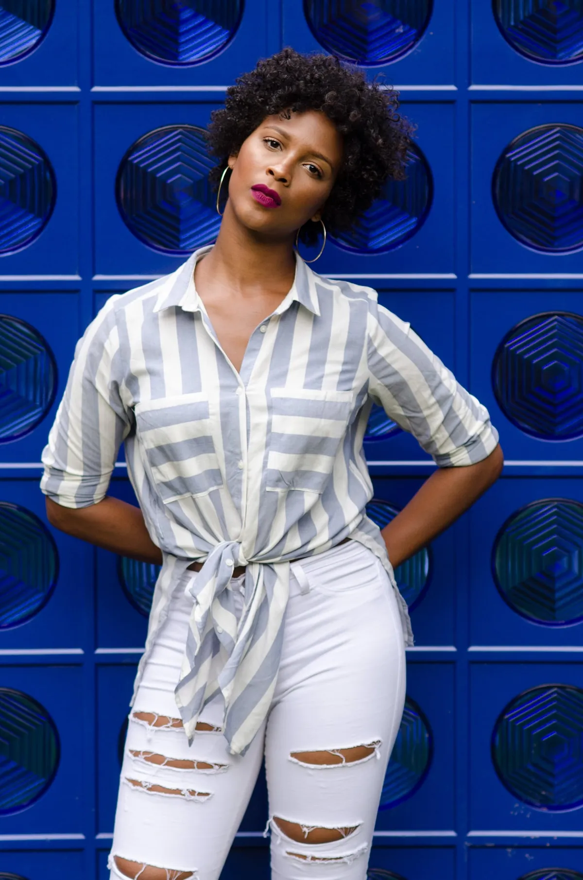A woman in a tied striped shirt and ripped white jeans poses against a bright blue patterned wall