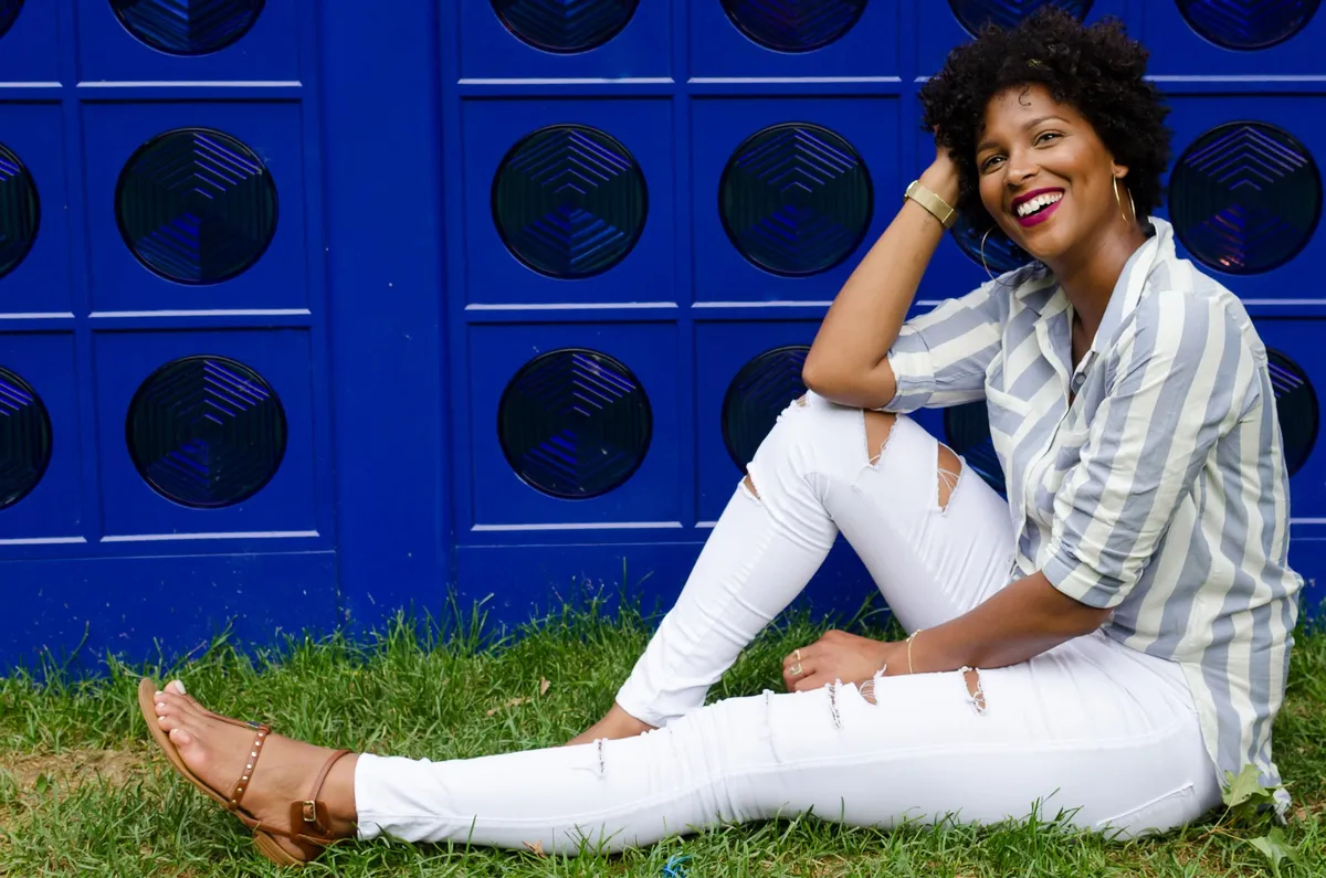 A woman sits smiling on the grass in ripped white jeans and sandals in front of a cobalt blue wall