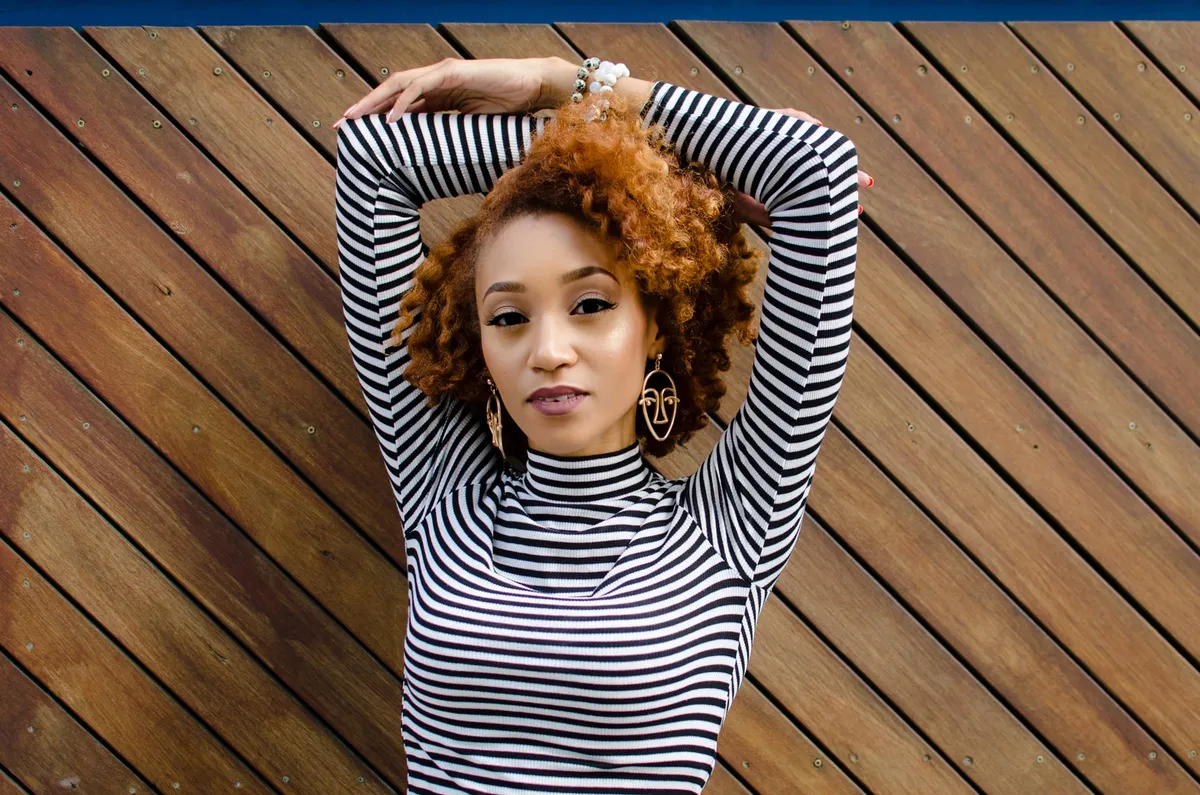 A woman with her arms raised overhead poses against a diagonal wooden wall