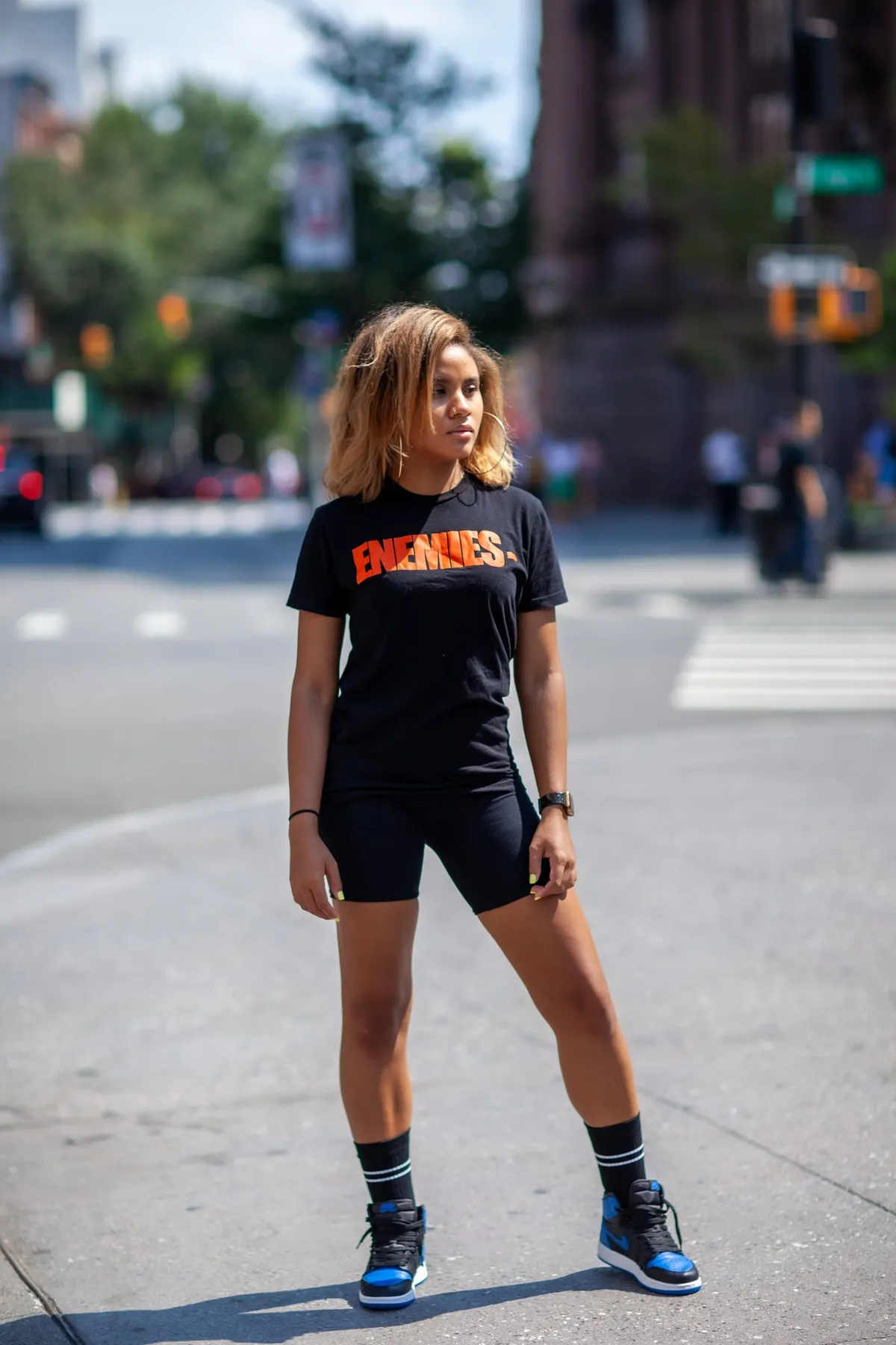 A woman in a black graphic tee and bike shorts stands on a city crosswalk