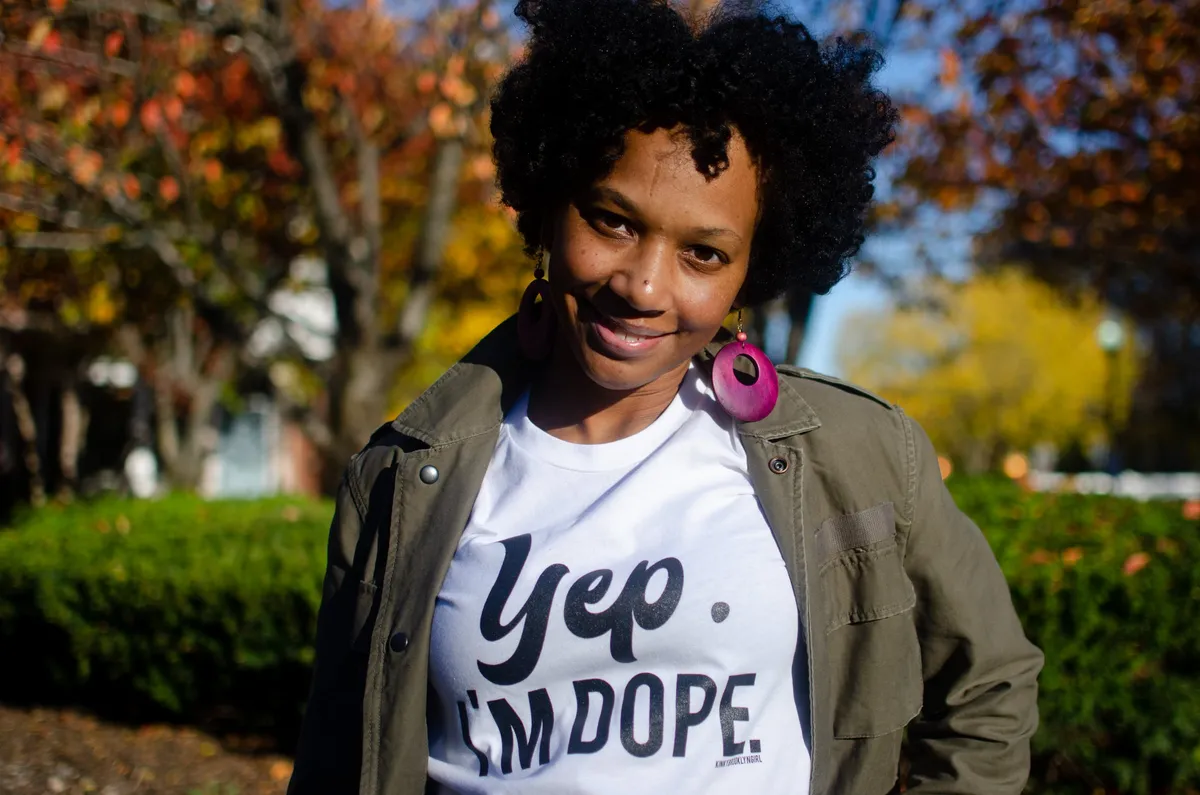 A woman in a "Yep, I'm Dope" t-shirt smiles in front of fall foliage