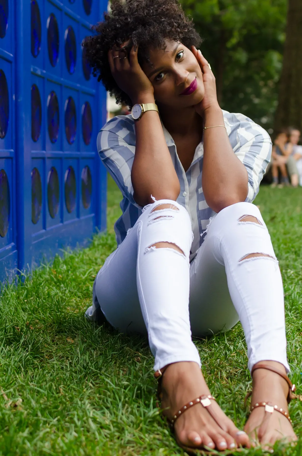 A woman sits on the grass with her hands in her hair beside a blue wall and green trees