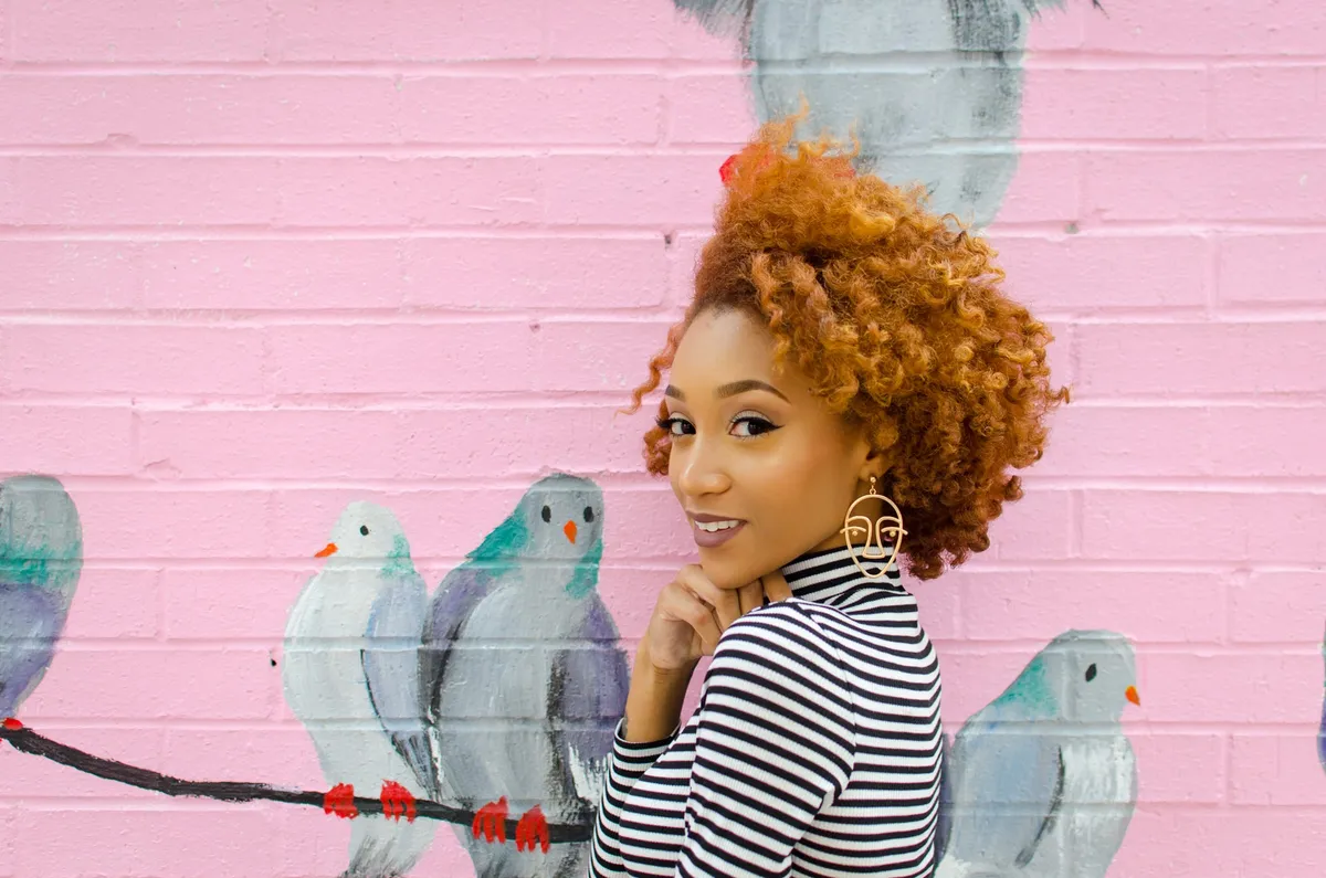 A woman in a striped turtleneck smiles beside a mural of pigeons on a pink wall