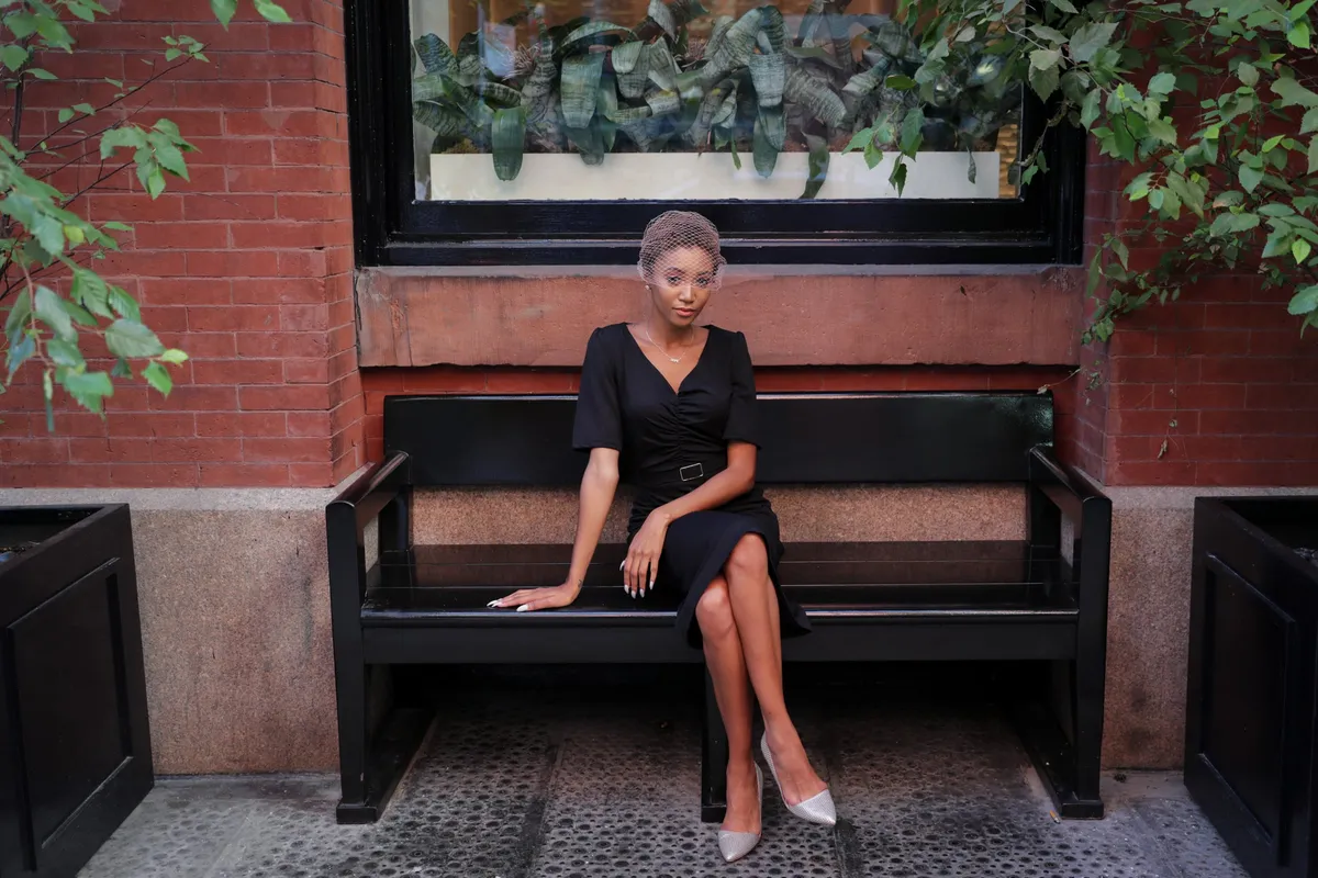 A woman in a black dress sits on a black bench beneath a window box
