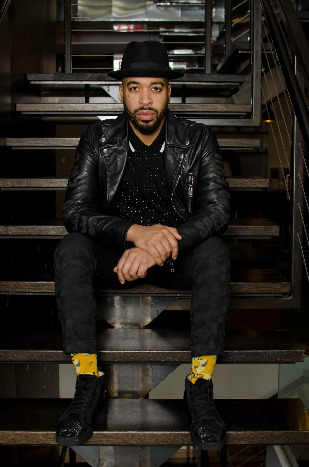 A man in a black fedora and leather jacket sits on a dark metal staircase wearing bright yellow socks