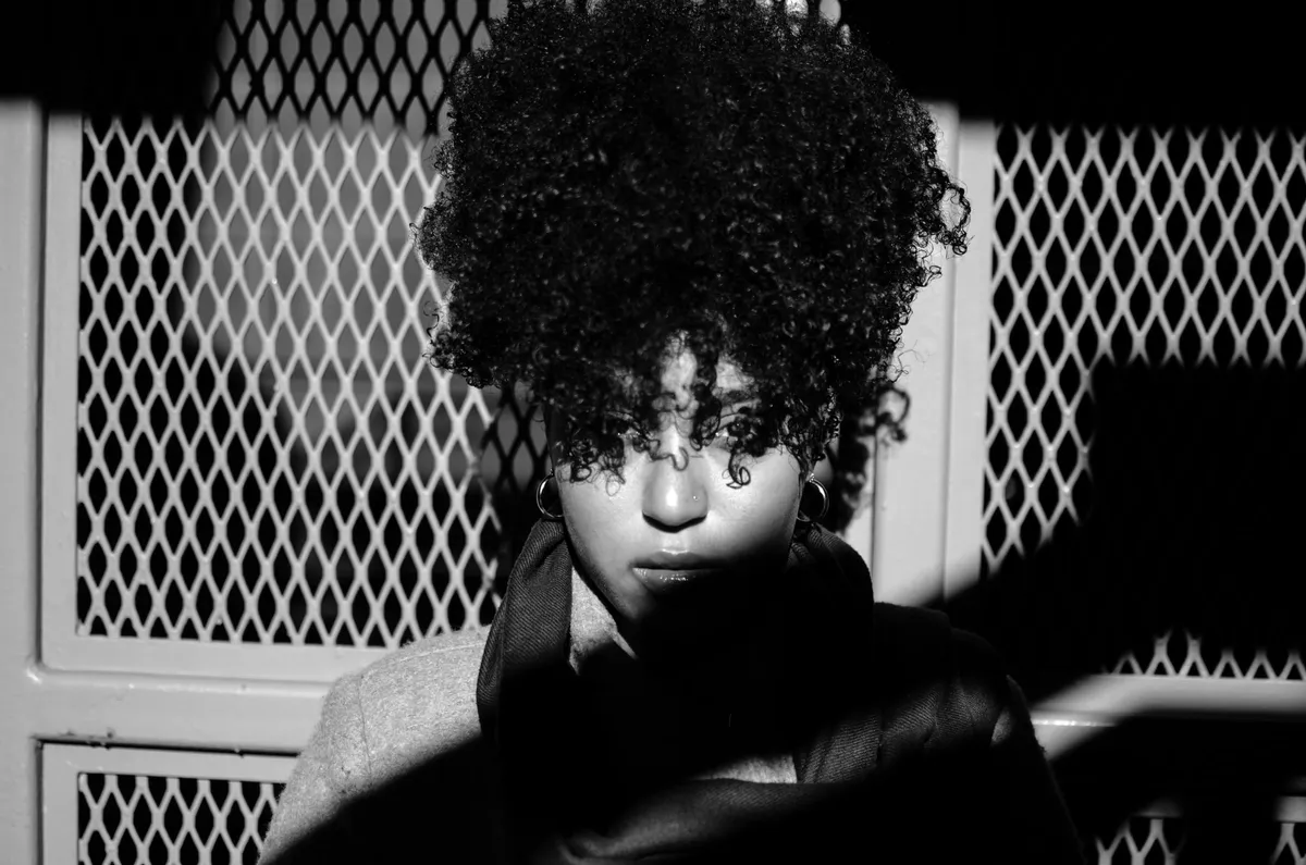 Black-and-white portrait of a woman with a full natural afro in dappled shade behind a lattice