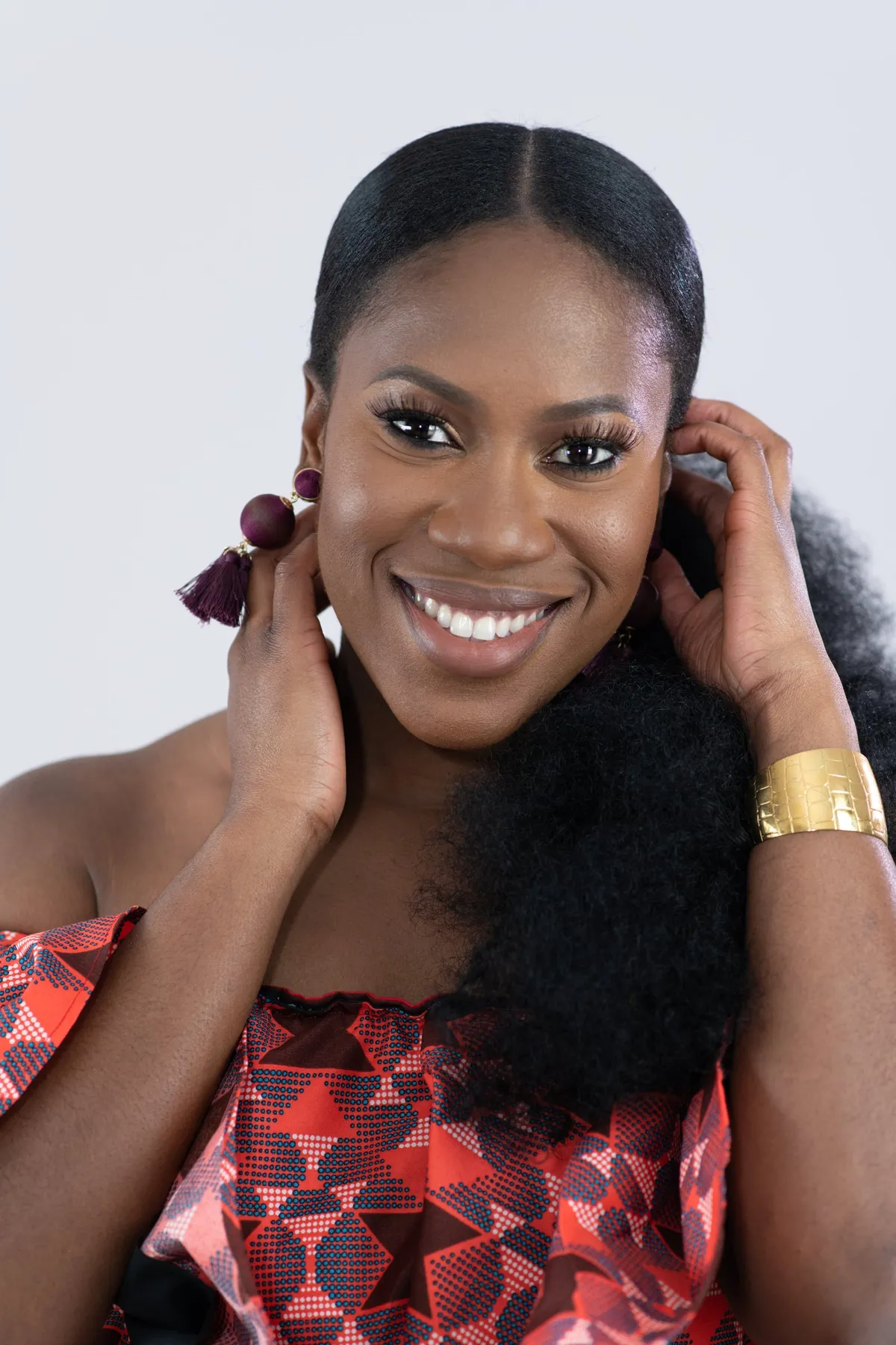 Studio portrait of a smiling woman with hands in her hair, wearing gold cuffs and a red patterned top