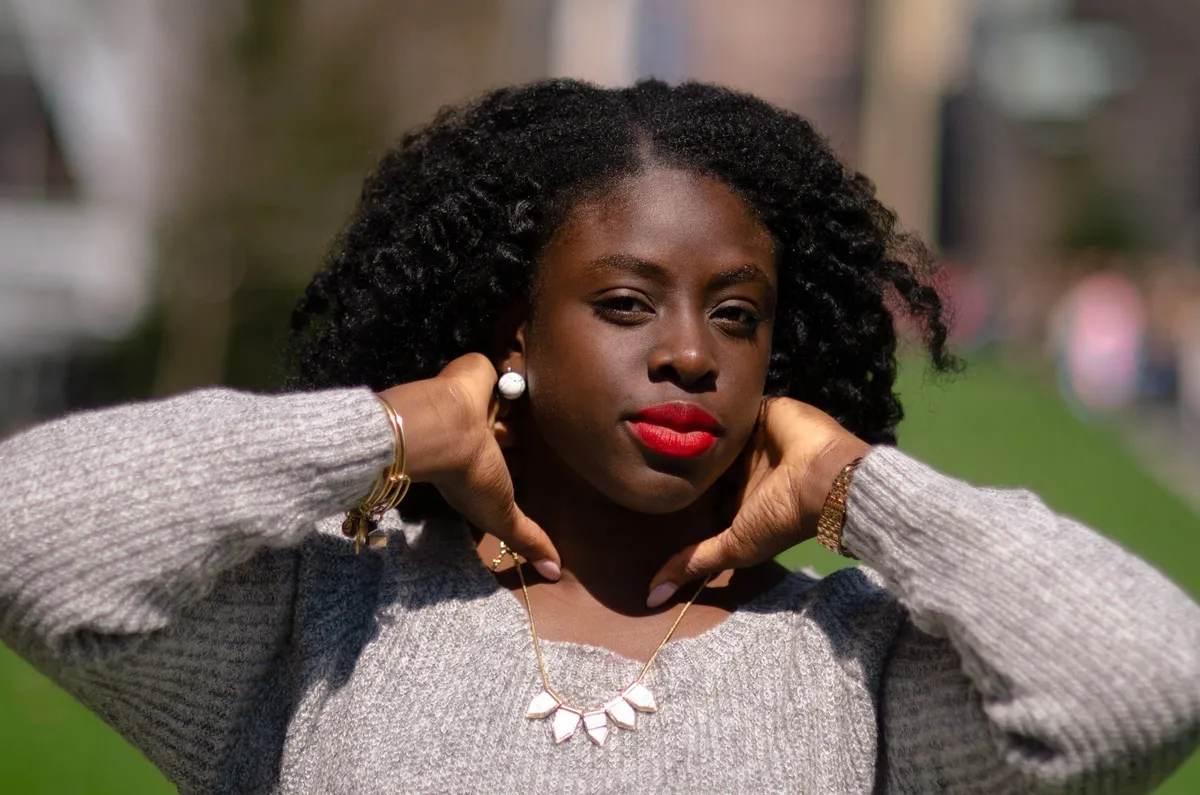 A woman with red lipstick and natural curls poses with her hands at her neck on a sunny lawn