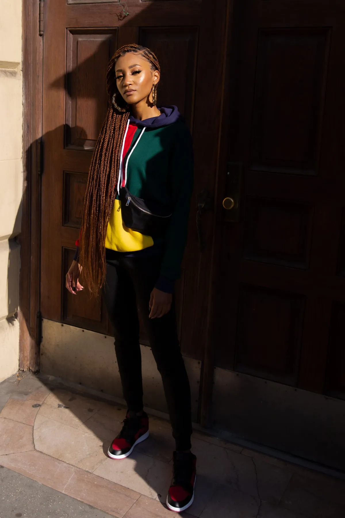 A woman with long auburn box braids in a color-block hoodie leans against a dark wooden doorway in afternoon sun