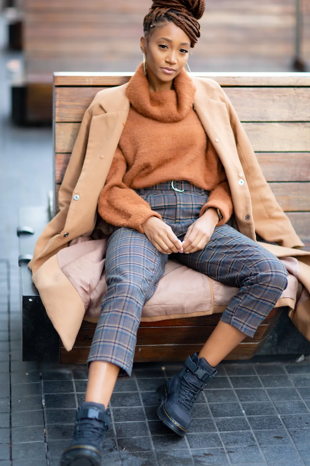 A woman in a camel coat and rust turtleneck sits on a wooden bench in plaid trousers and boots