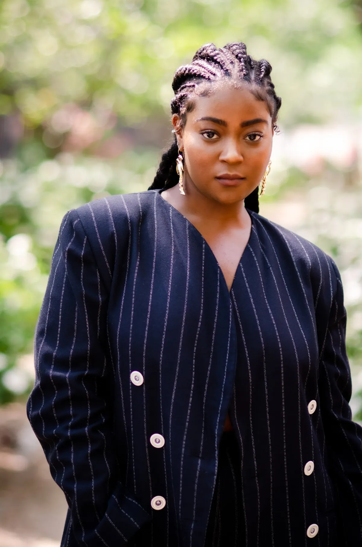 A woman with braided hair in a navy pinstripe blazer stands in a garden