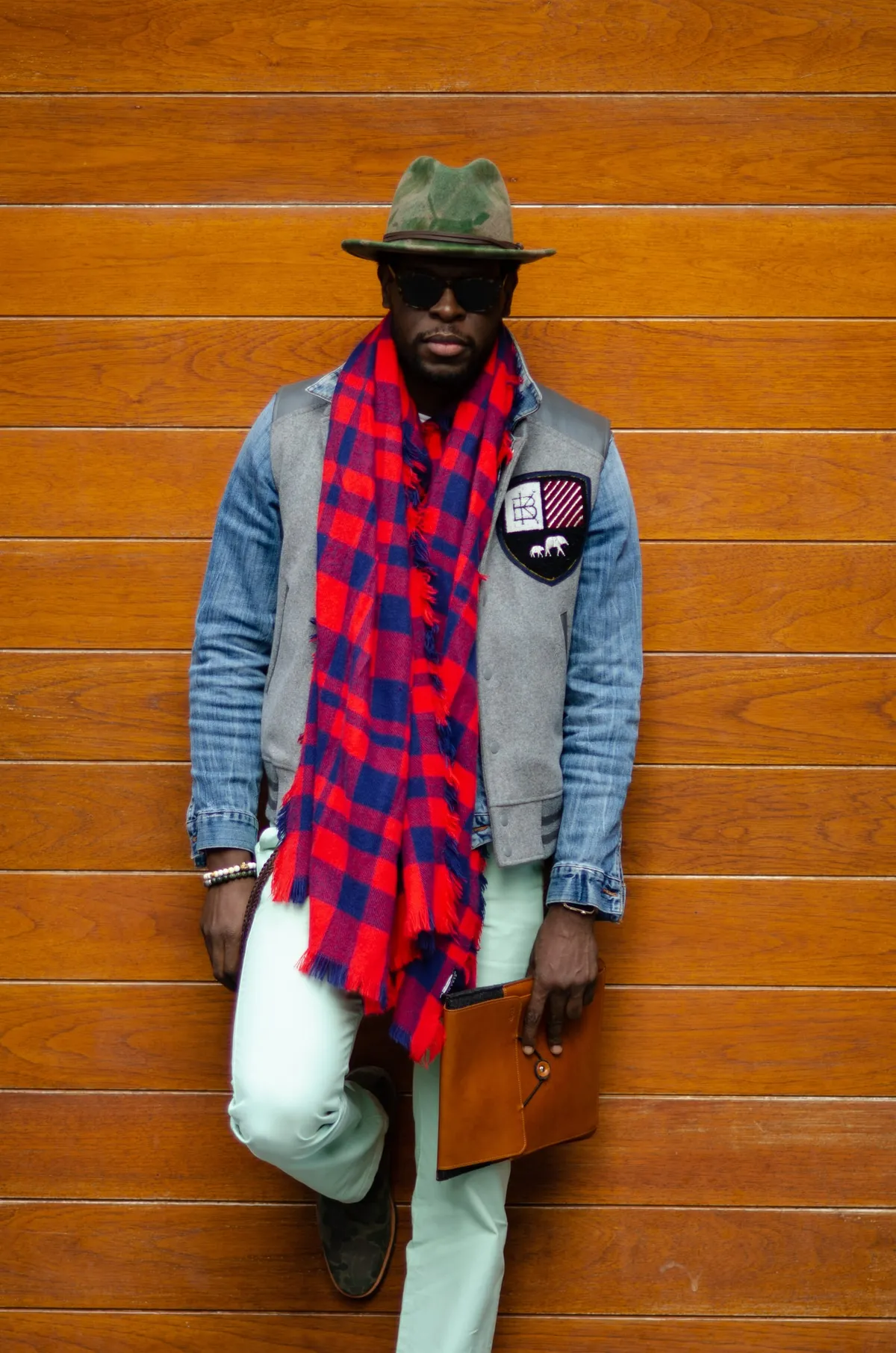 A man in a green fedora, denim jacket and red plaid scarf leans against an orange wooden wall holding a leather folio