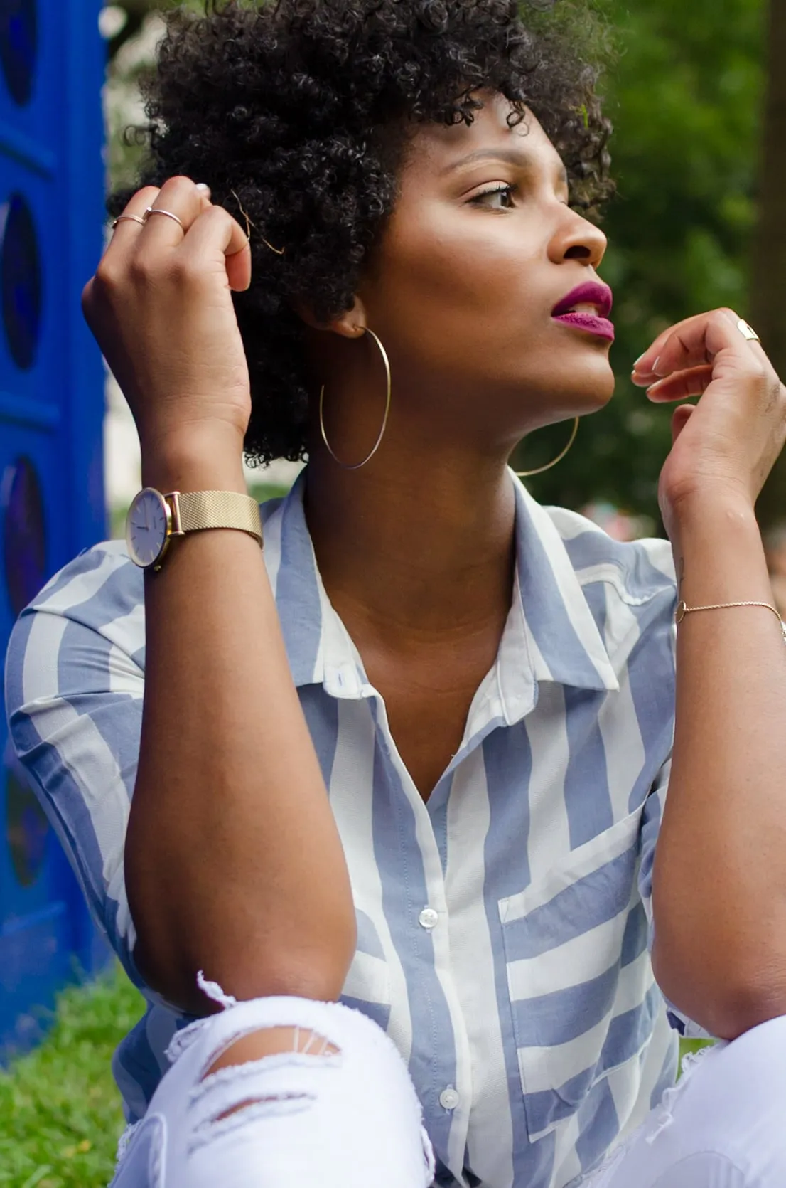 A woman rests her hands near her face, gold hoops and a watch catching the light, against a blue wall