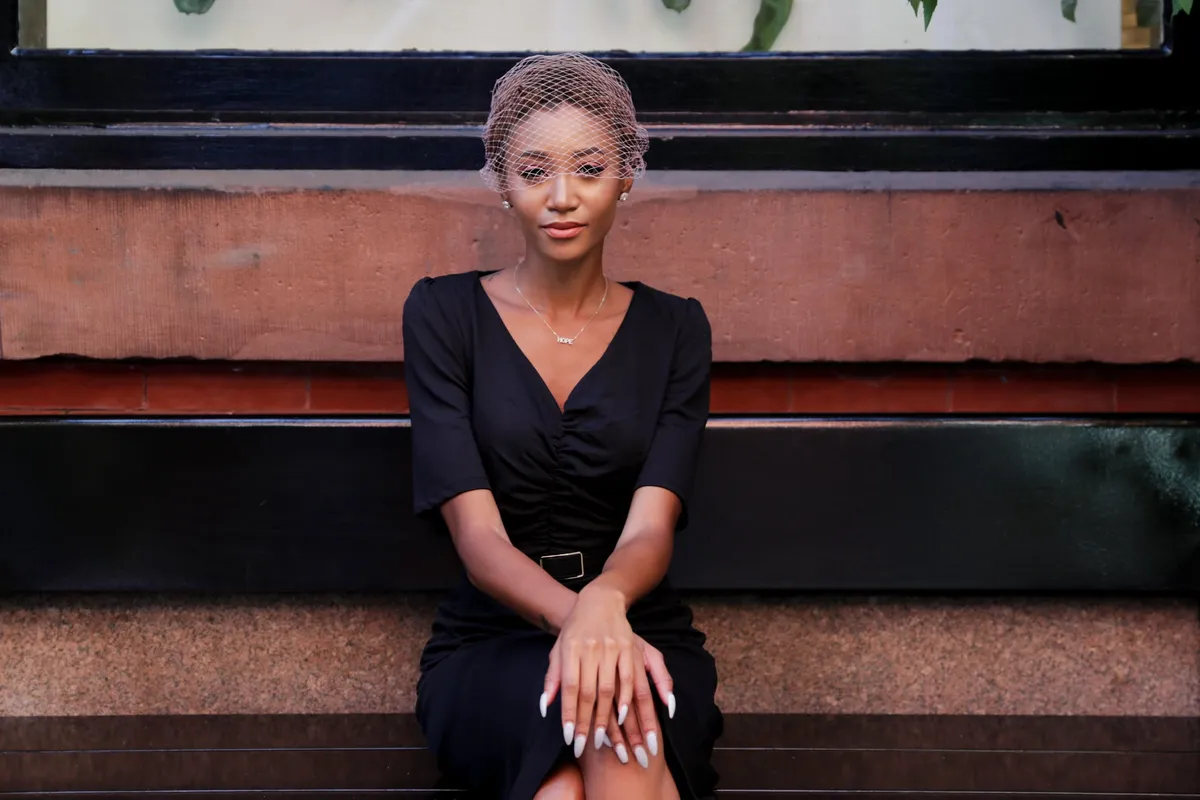 A woman sits on a storefront ledge in a black dress, hands folded
