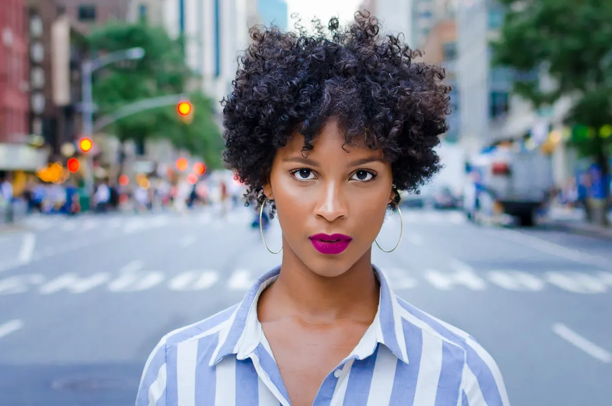 A woman with magenta lips stands in the middle of a city avenue, traffic and trees blurred behind her