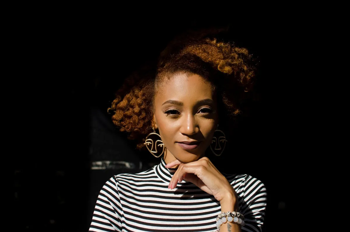A woman rests her chin on her hand in a pool of light against a black background
