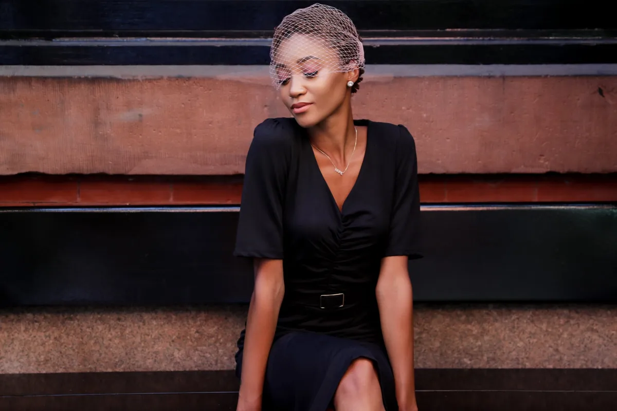 A woman with her eyes lowered sits on a storefront ledge in a black dress