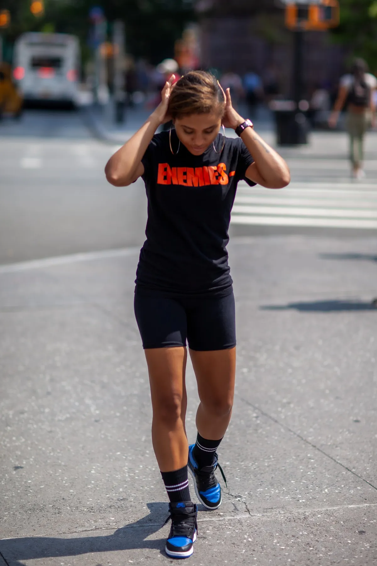 A woman walks toward the camera on a city street, both hands in her hair