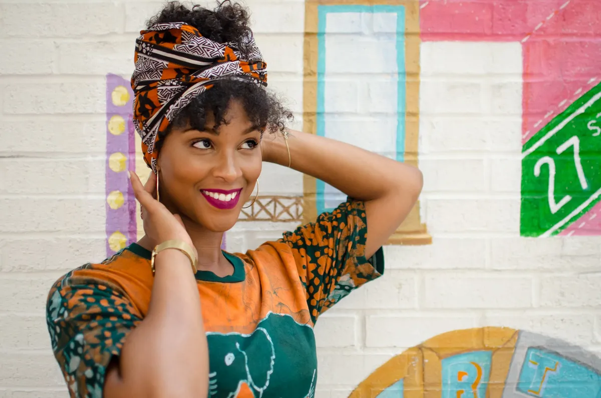 A woman in a patterned head wrap smiles with hands raised to her head in front of a colorful mural