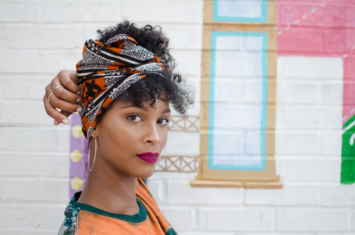 A woman in an orange and black head wrap glances sideways in front of a painted brick wall