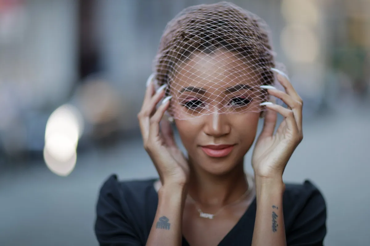 A woman in a birdcage veil frames her face with her hands on a softly blurred street