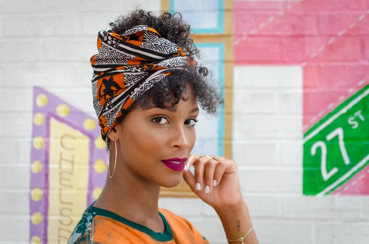 A woman in a head wrap rests her hand by her chin beside a mural with a West 27th Street sign
