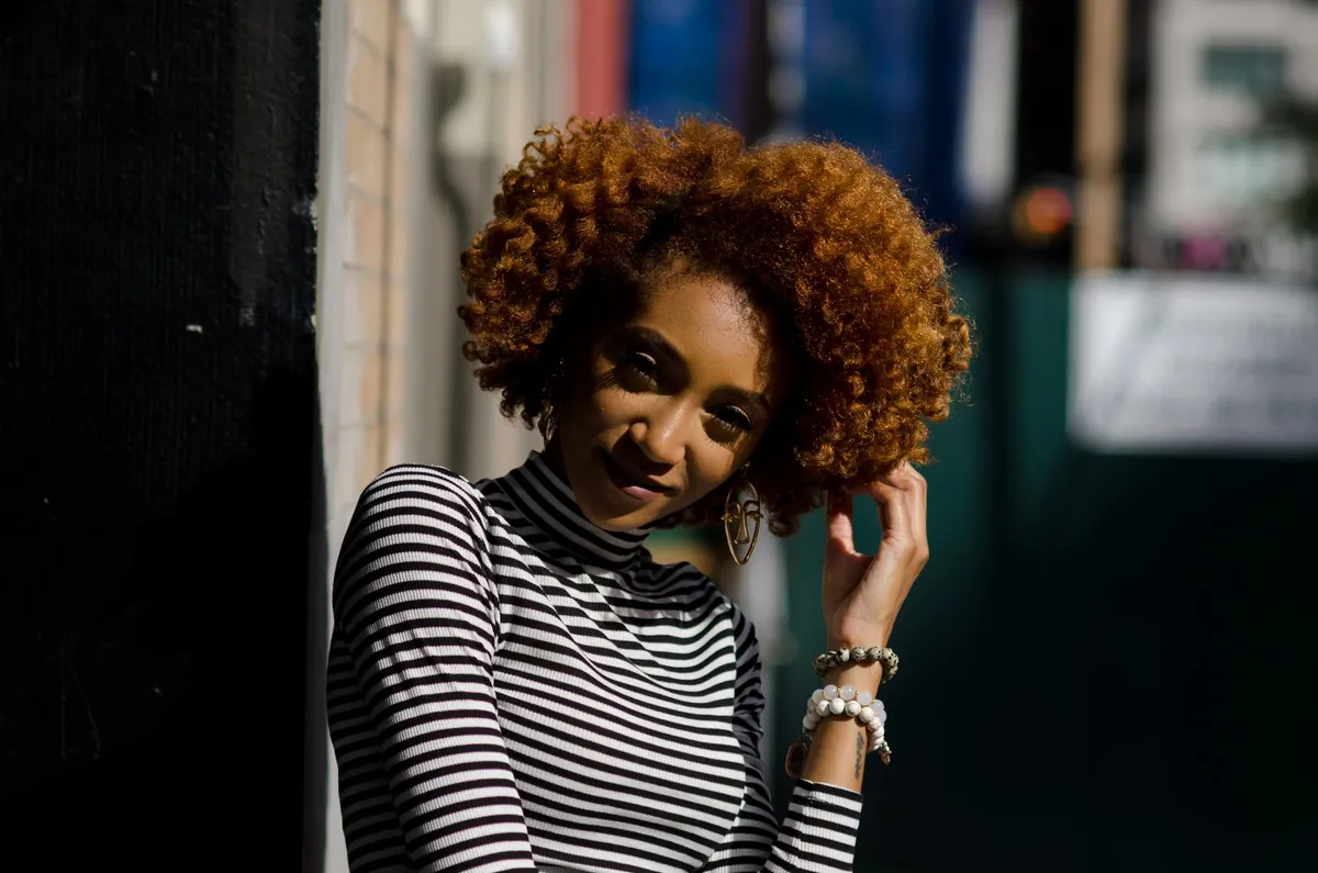 A woman with curly auburn hair smiles softly in a shaded doorway
