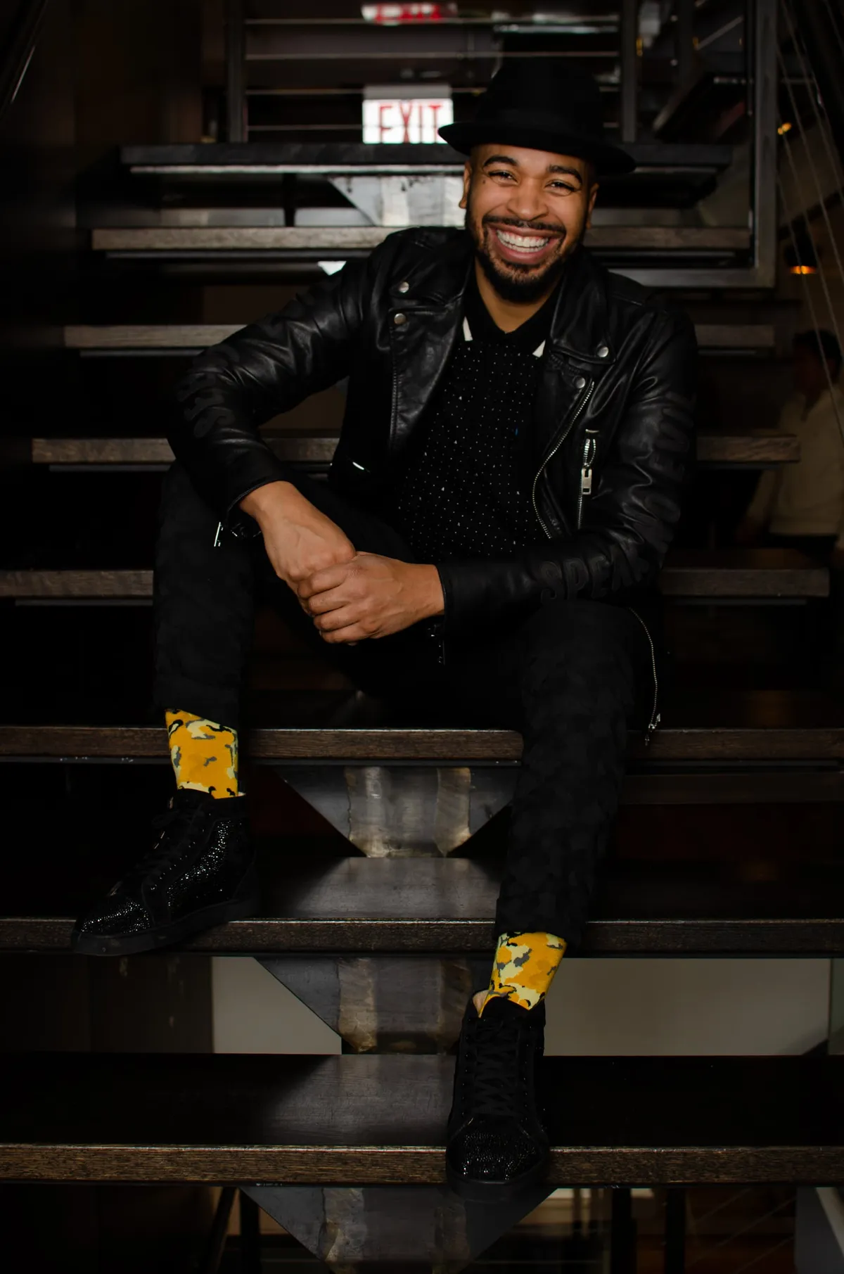 A man in a fedora and leather jacket laughs while seated on industrial stairs