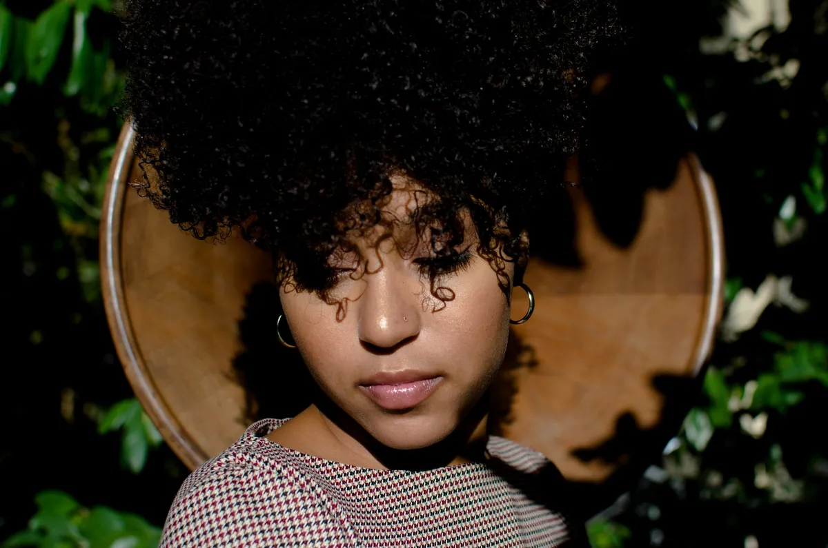 A woman with voluminous curls closes her eyes in front of a round wooden panel framed by leaves