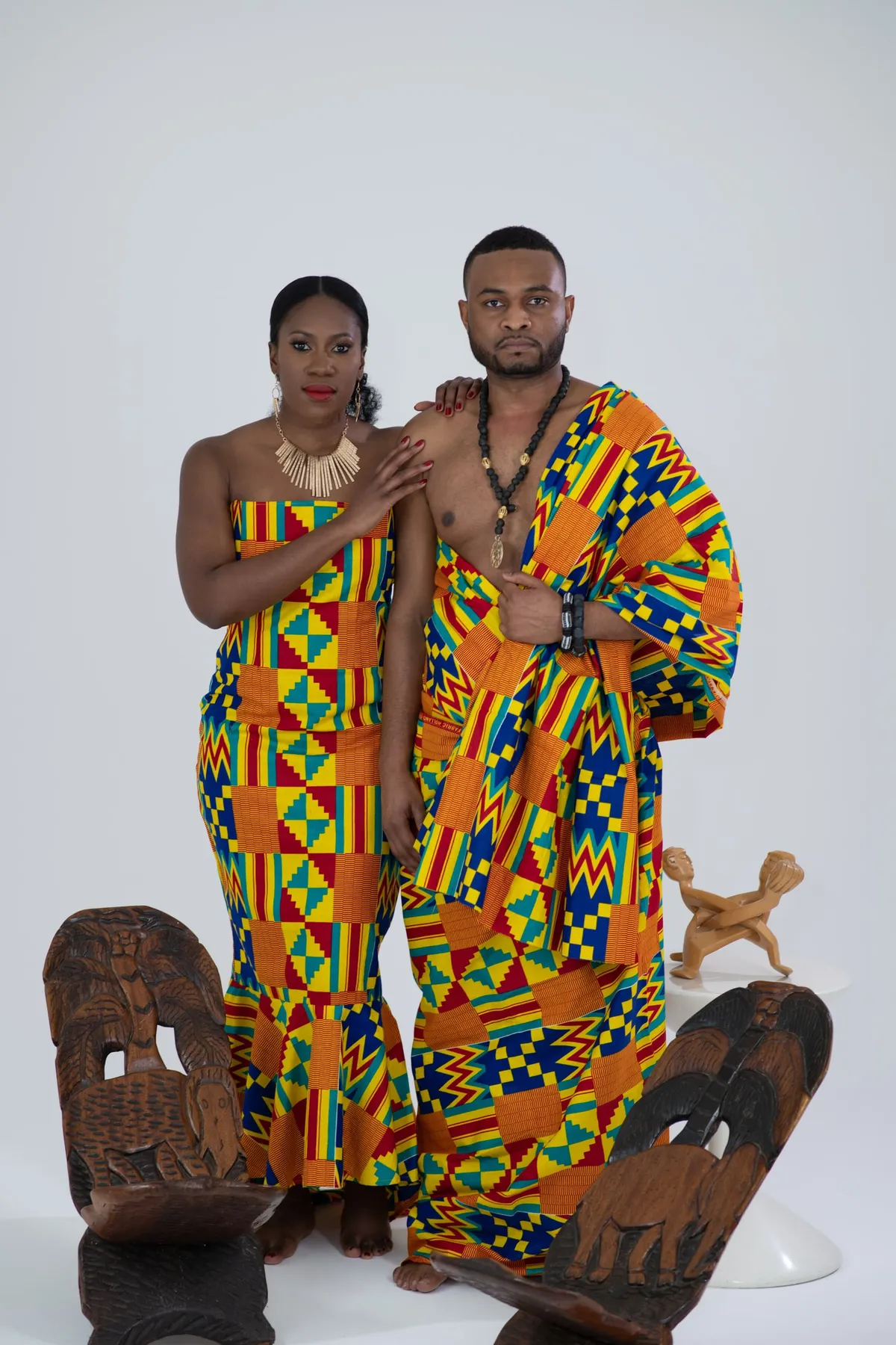 A couple in matching kente cloth outfits pose in the studio between carved wooden chairs